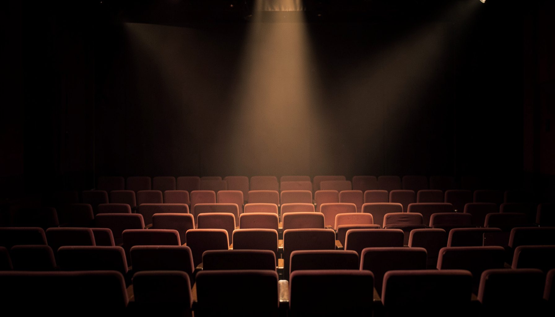 Warm-toned photograph of empty theater seats lit by a soft amber spotlight on a dim stage, muted rose and charcoal tones.